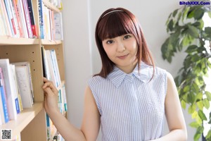 A woman in a blue and white shirt is standing in front of a bookshelf.