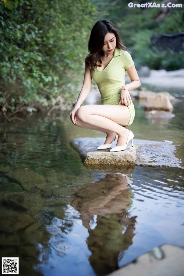 A woman in a green dress sitting on a rock in the water.